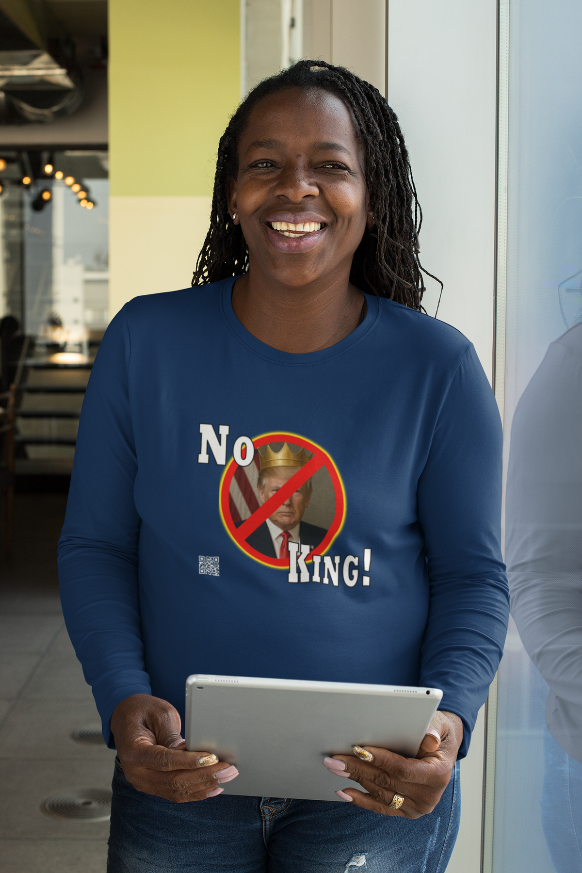 A smiling woman wearing a navy blue long-sleeve t-shirt with a 'No King!' graphic print, holding a tablet in a brightly lit indoor setting.