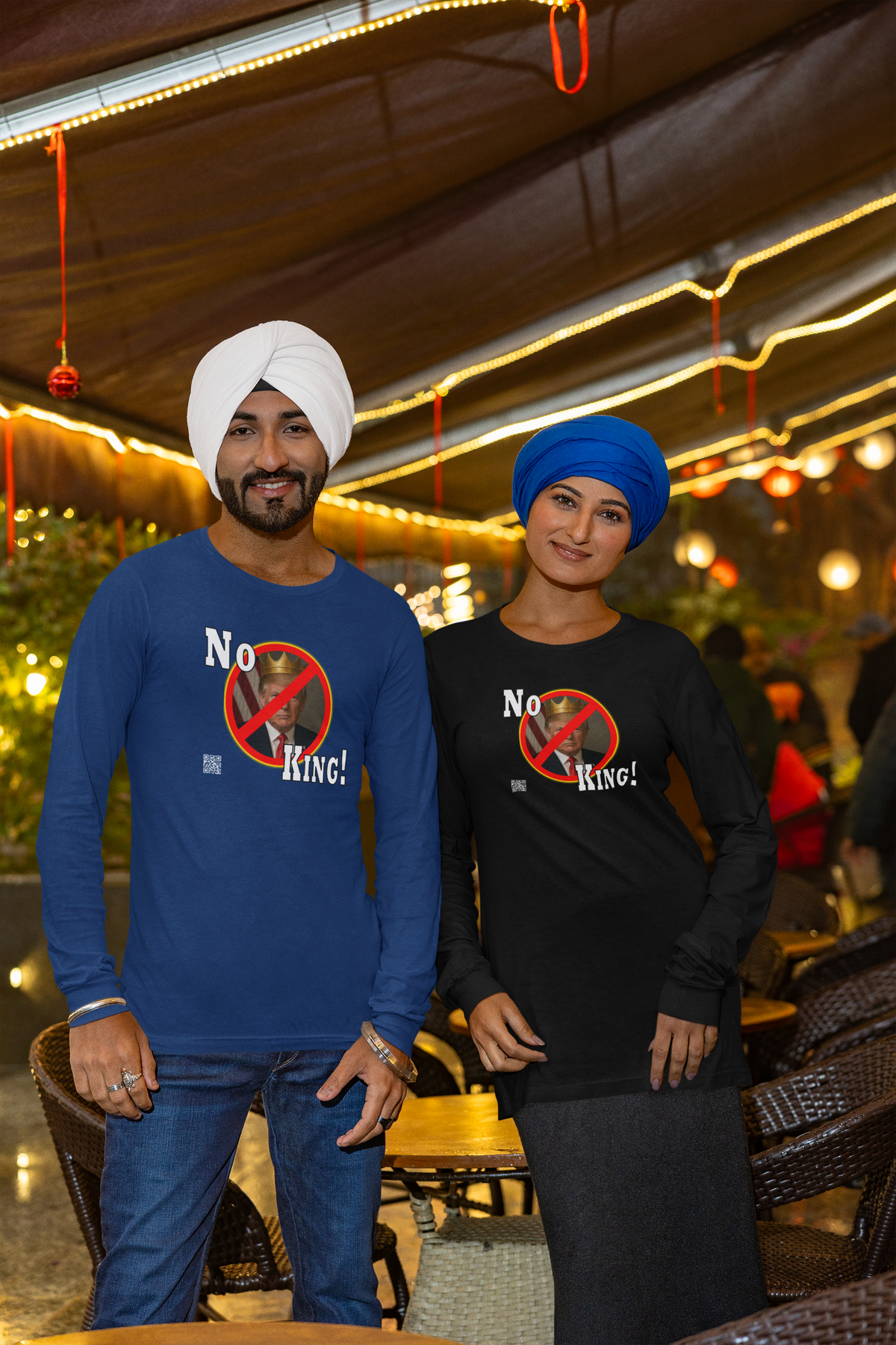 A man and a woman standing side-by-side at an outdoor night market or restaurant, wearing matching long-sleeve graphic t-shirts. The man wears a blue turban and a blue shirt with a graphic of a bear inside a prohibition sign with the text 'No King!'.