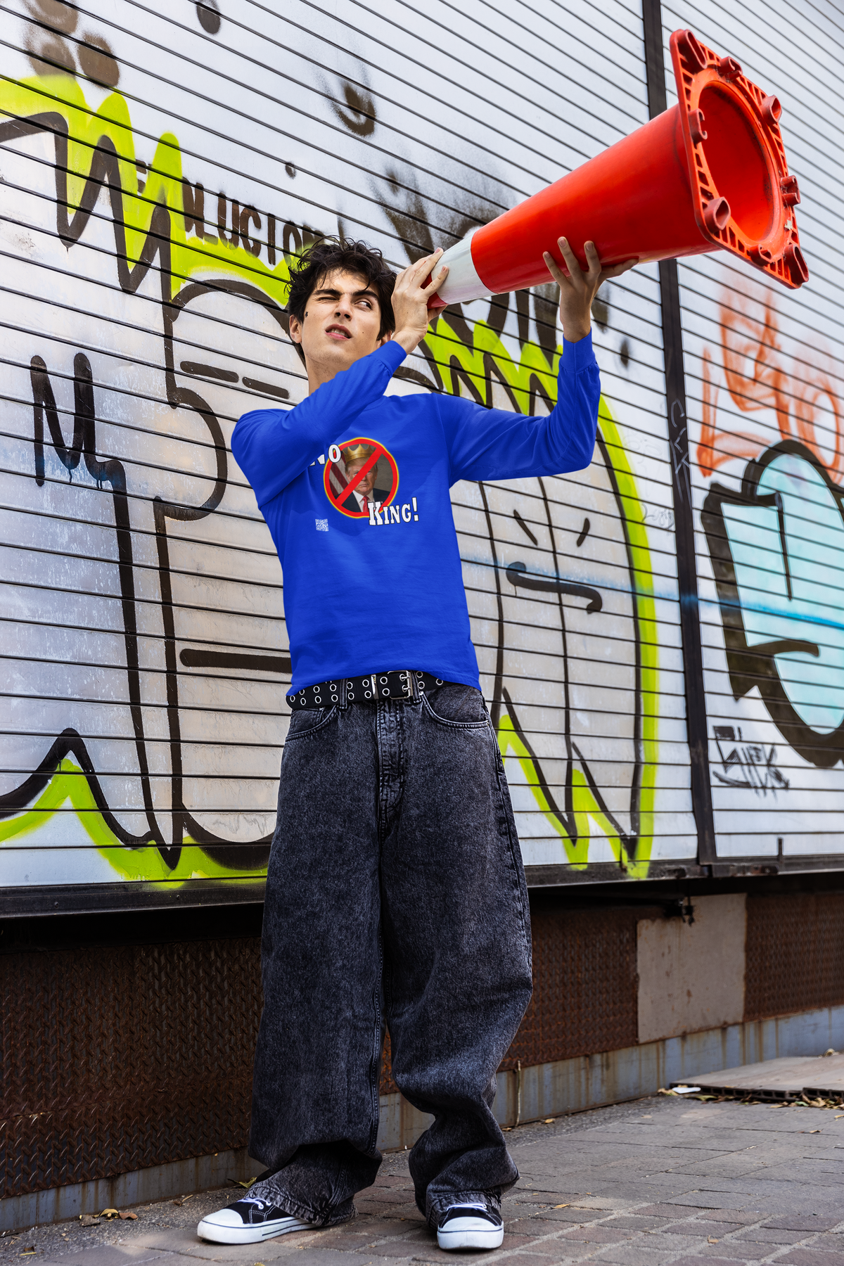 A person stands in front of a graffiti-covered metal shutter, holding a red traffic cone over their head like a megaphone. They are wearing a bright blue long-sleeve t-shirt with a graphic on the chest, loose dark gray jeans, and black-and-white sneakers.